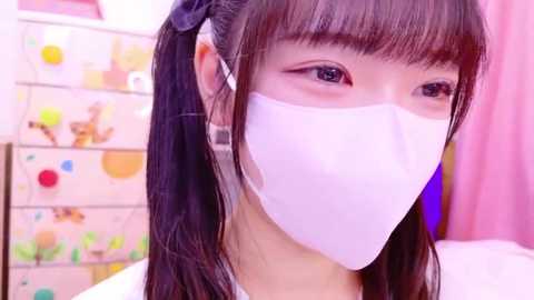 Media: Video of an East Asian woman with long black hair in pigtails, wearing a white face mask, in a colorful, playful room with pastel-colored furniture and decorations.