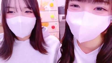 Media: Video of two young Asian women with long black hair, wearing white surgical masks, standing in a bright, colorful room with pastel-colored shelves in the background.