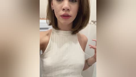 Media: Video of a young, fair-skinned woman with short, straight brown hair, wearing a white sleeveless, high-neck top. She stands in a bathroom, holding a small item. The image is slightly blurred.