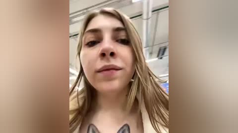 Media: Video of a young Caucasian woman with wet, blonde hair, wearing a white top with a black dog tattoo on her chest, standing in a modern, white-tiled bathroom with visible pipes and a showerhead.