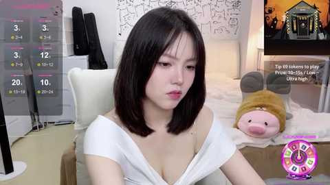 Media: Video of a young East Asian woman with shoulder-length black hair, wearing a white off-shoulder top, sitting in a room with a plush toy, TV, and digital clock.