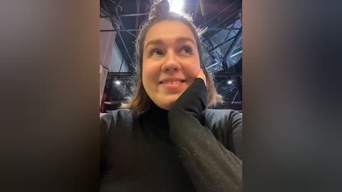 Media: A video of a young woman with light skin and brown hair, smiling and holding a large, dark object in her hand, taken indoors in a dimly lit industrial space with exposed beams and metal structures.