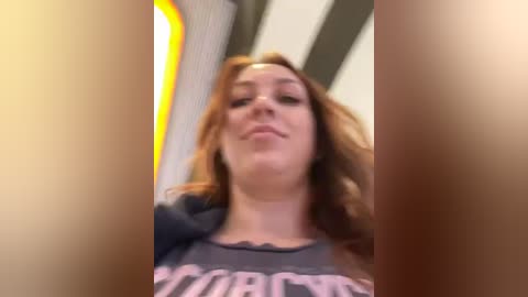 Media: Video of a woman with long, wavy red hair, wearing a dark grey T-shirt with a pink design. She has a fair complexion and a confident, slightly playful expression. The background is blurry, suggesting a low-angle perspective.