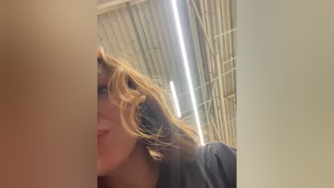 Media: A close-up video of a smiling woman with light brown hair, wearing a dark shirt, taken in a bathroom with a tiled ceiling and fluorescent lighting.