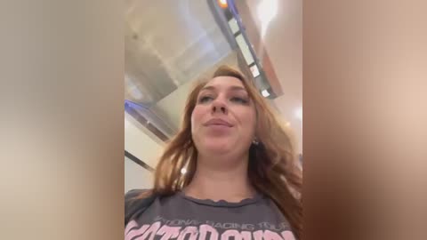 Media: A video of a smiling, light-skinned woman with long red hair, wearing a dark T-shirt with a colorful graphic design, standing indoors near a staircase.