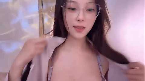 Media: Video of an Asian woman with fair skin, long black hair, and glasses, wearing a white shirt, playfully pulling her hair, with a blurred background.