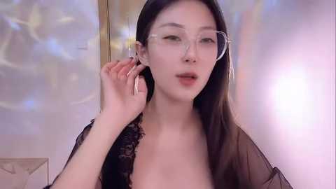 Media: A video of an East Asian woman with long, straight black hair, fair skin, and large hoop earrings, wearing a sheer black top, adjusting her glasses. The background features soft, pastel-colored lights.