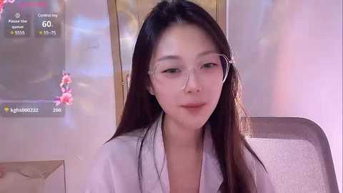 Media: A video of an Asian woman with long dark hair and glasses, wearing a white robe, sitting in a dimly lit room with a pink flowered wall and a digital clock.