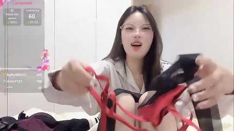 Media: Video of a smiling East Asian woman with long black hair, fair skin, wearing glasses, holding red lingerie, in a modern bedroom with white walls and a calendar.