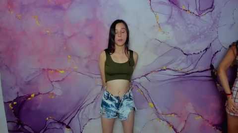 Media: A video shows a young woman with long dark hair, wearing a green crop top and blue tie-dye shorts, standing against a vibrant, abstract purple and pink background.