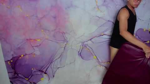 Media: Video of a young, light-skinned man with glasses, wearing a black sleeveless shirt, standing against a vibrant, abstract, marble-like wall mural with purple, pink, and white hues.