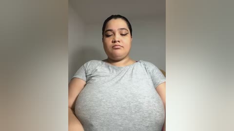 Media: Video of a plus-sized, light-skinned woman with dark hair, wearing a grey t-shirt, standing against a plain beige wall, eyes closed, looking serene.