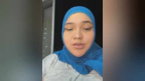 Media: A video of a young woman with medium skin tone, wearing a bright blue hijab and a grey shirt, standing in a dimly lit room with a black background.