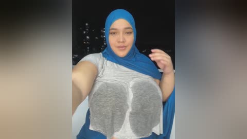 Media: Video of a plus-size woman with a blue hijab, gray top, and blue cape, posing in front of a dark cityscape at night.