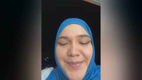 Media: Video of a smiling young woman with light brown skin, wearing a blue hijab and makeup, set against a dark background.