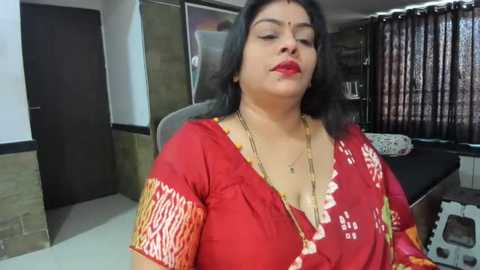 Media: Video of a middle-aged woman with long black hair, wearing a red sari with white floral patterns, seated in an indoor setting.