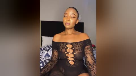 Media: Video of a black woman with short hair, wearing a black, lace bodysuit with an off-the-shoulder neckline and intricate cutouts, sitting on a bed with patterned bedding in a modern, minimalist bedroom.