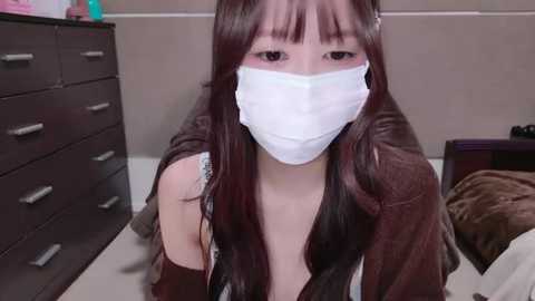 Media: Video of a young Asian woman with long, dark hair, wearing a white face mask, brown top, and sitting on a bed in a beige room with a dark dresser.