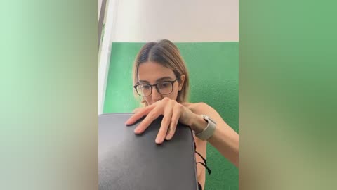 Media: Video of a young woman with long brown hair and glasses, wearing a black tank top, standing in a room with green and white walls, using a laptop.