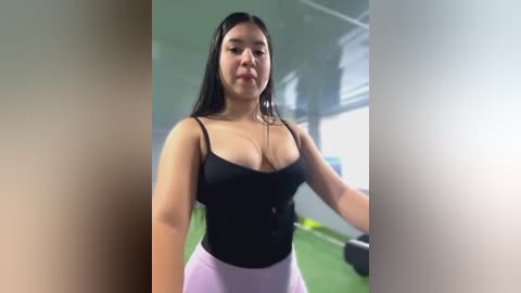 Media: Video of a Latina woman with long black hair, medium build, wearing a black tank top and pink leggings, standing in a gym with green flooring and equipment in the background.