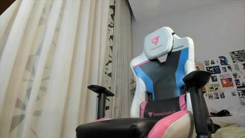 Media: Video of a pink and white Razer gaming chair with adjustable armrests in a cluttered bedroom with a bed, white curtains, and a wall covered in polaroid photos.