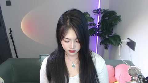 Media: Video of a young Asian woman with long black hair, wearing a white top, and a green necklace. She sits on a green couch with pink cushions, surrounded by a green screen and a potted plant.