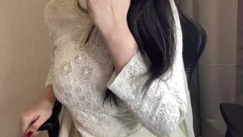 Media: A video of a woman with long, straight black hair, wearing a beige lace dress, sitting in a chair with a gray curtain in the background.