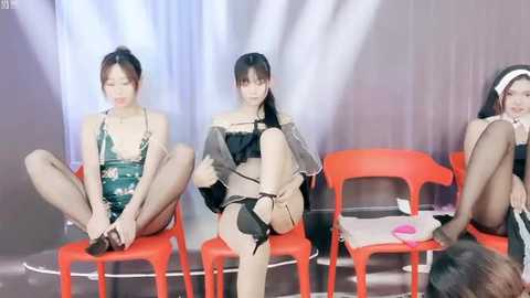 Media: Video of three Asian women in provocative lingerie, sitting on bright orange plastic chairs against a white backdrop, legs crossed, wearing sheer stockings and high heels.