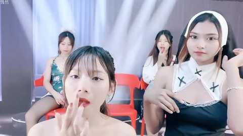 Media: A video of four Asian women in a modern studio setting. Three women in revealing black and white nun costumes, one with a red chair, another with a solemn expression, and the fourth in the foreground.