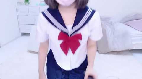 Media: Video of a young woman with shoulder-length brown hair, wearing a Sailor Moon school uniform with a large red heart on the chest, standing in a minimalist bedroom with white walls, a bed, and dresser.