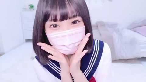 Media: Video of an Asian woman with straight black hair and bangs, wearing a white surgical mask and a sailor school uniform, with hands placed gently on her cheeks. Background features a white bed and white walls.