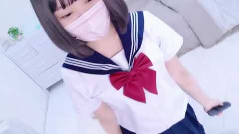 Media: A video of an Asian woman in a white sailor uniform with a red bow, a pink face mask, and a gray controller, standing in a minimalist, light-colored room.