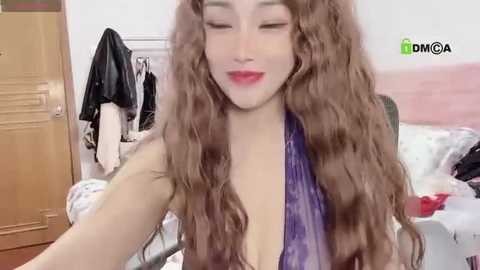 Media: Video of an East Asian woman with long, wavy brown hair, wearing a purple dress, smiling with closed eyes in a messy bedroom.