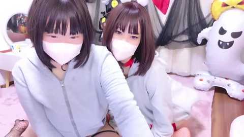 Media: Video of two young Asian women with straight black hair, wearing light blue jackets, white masks, and black fishnet stockings, seated on a pink carpet, surrounded by Halloween decorations.