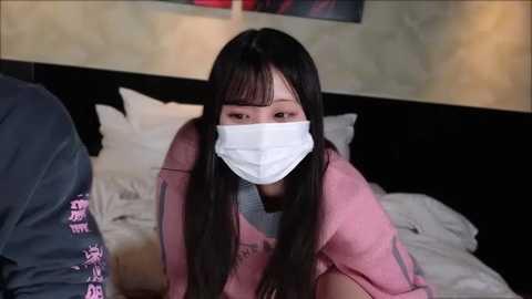 Media: Video of an Asian woman with long black hair and a white surgical mask, wearing a pink robe, kneeling on a bed with white linens in a dimly lit room.