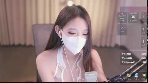 Media: A video of a young Asian woman with long, straight black hair, wearing a white face mask and headphones, sitting at a desk in a dimly lit room.