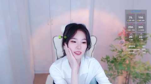 Media: A video of a smiling East Asian woman with black hair, wearing a white shirt, sitting in a chair, with a green hair clip. The background shows a white wall, a plant, and a virtual display of social media stats.