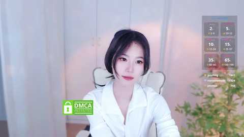 Media: Video of an East Asian woman with short black hair, wearing a white blouse, seated in a chair, against a softly lit, white backdrop. The image features a virtual thermometer overlay.