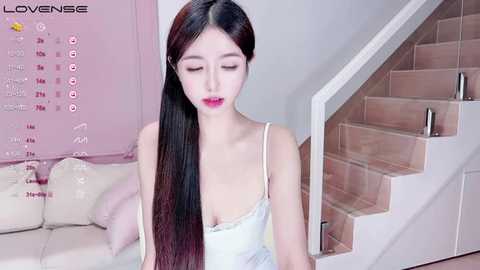 Media: Video of an East Asian woman with long black hair, fair skin, and pink lipstick, wearing a white spaghetti-strap camisole, sitting on a beige couch, next to a wooden staircase.