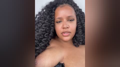 Media: Video of a young Black woman with a light brown complexion, large natural breasts, and curly black hair, wearing a black strapless top, against a blurred background.