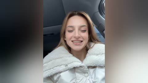 Media: Video of a young Caucasian woman with light brown hair, wearing a white fuzzy jacket, smiling with eyes closed inside a car.