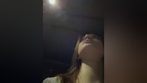 Media: A video captures a young woman with straight, light brown hair and fair skin, gazing upwards. The dimly lit room has a lens flare above her head, enhancing the moody atmosphere.