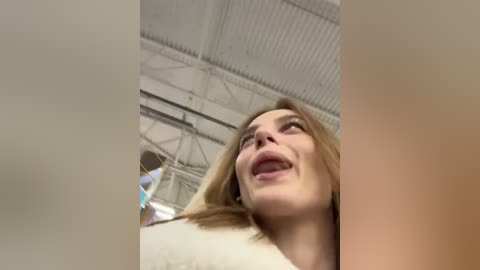 Media: A video captures a blonde woman with a joyful expression, seen from a low angle, against a white, industrial ceiling with metal beams. Her light skin and white clothing contrast with the background.