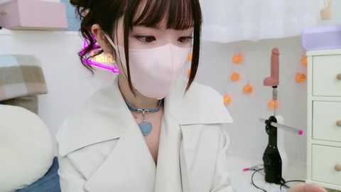 Media: Video of a young Asian woman with fair skin and dark hair, wearing a white coat, face mask, and purple earrings, in a sterile, minimalist room with white walls, a toy, and a dildo.