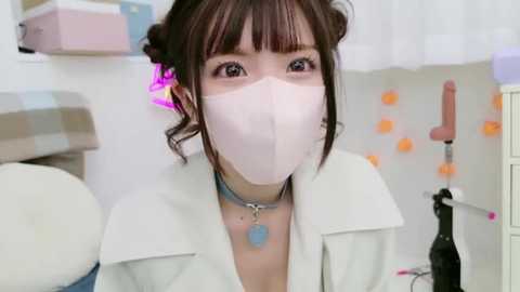 Media: A video of an Asian woman with dark hair styled in pigtails, wearing a white lab coat, pink face mask, and blue dog tag necklace. Background features medical equipment and white walls.