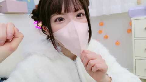 Media: Video of an Asian woman with dark hair, wearing a pink face mask, white fur coat, and white gloves, playfully holding a pink feather duster in a bright, clean room with white walls and orange decor.