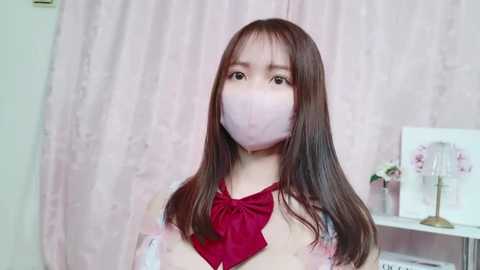 Media: Video of an Asian woman with long brown hair, wearing a red bow and pink face mask, standing in front of a pink, textured curtain.