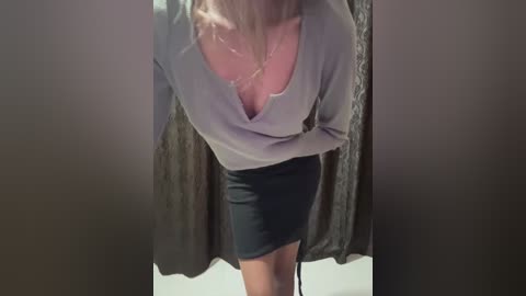 Media: Video of a woman with medium skin tone, wearing a low-cut, grey sweater revealing a pink bra, black mini skirt, and standing in front of a dark curtain.