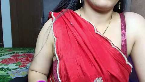 Media: Video of a woman in a red sari with gold embroidery, wearing a gold necklace and dangling earrings, standing in a room with a green floral-patterned rug and wooden furniture.