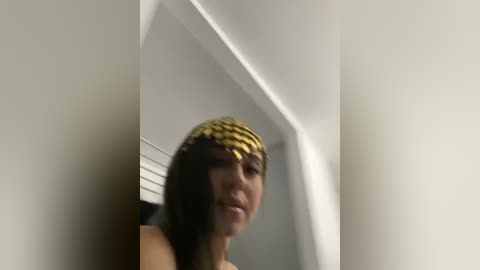 Media: Video of a young woman with straight black hair adorned with a golden headband, standing in a dimly lit room with white walls and a window partially visible in the background.